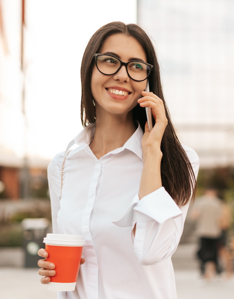 Mujer sonriendo mientras sostiene un café y habla por teléfono en la calle