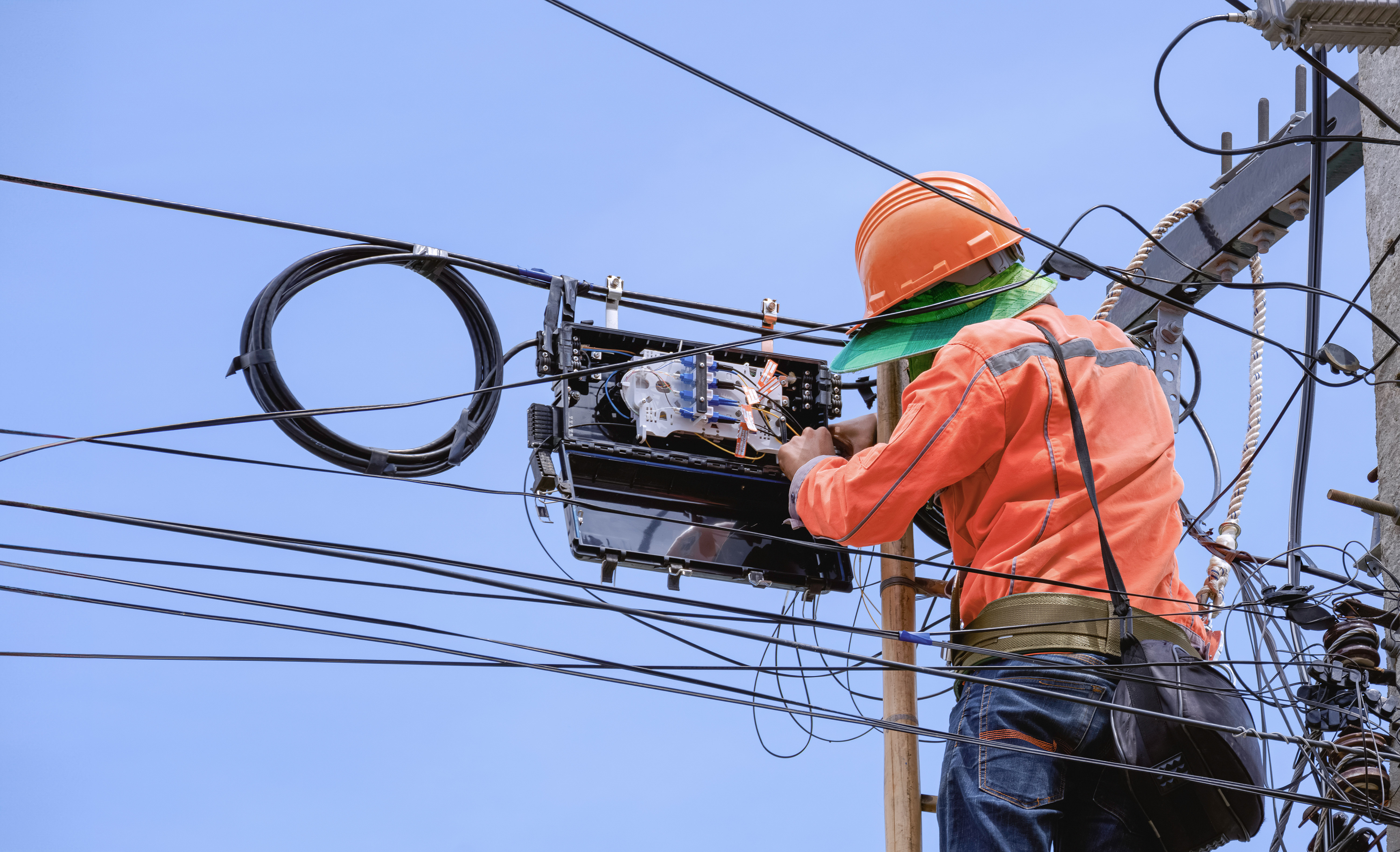 Técnico con casco naranja subido a un poste manipulando una caja de empalme de fibra óptica entre cables aéreos.