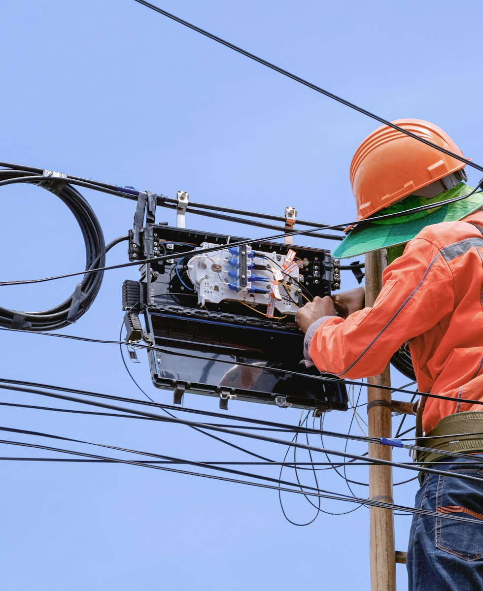 Técnico con casco naranja subido a un poste manipulando una caja de empalme de fibra óptica entre cables aéreos.
