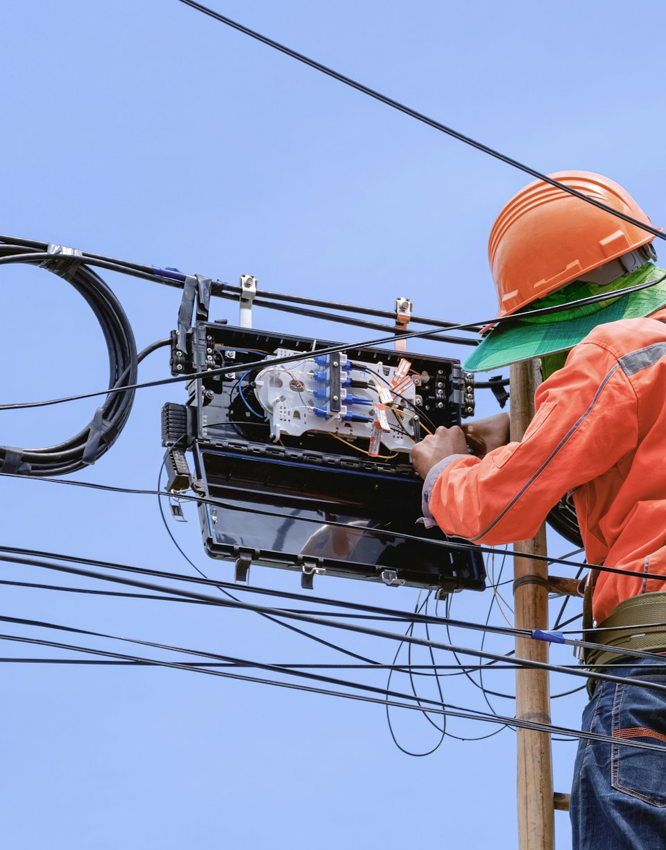 Técnico con casco naranja subido a un poste manipulando una caja de empalme de fibra óptica entre cables aéreos.